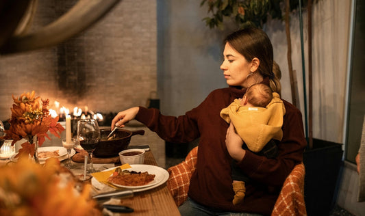 Photo of a peaceful new mother sitting at a dining table, holding her baby in her arms while eating a balanced, nourishing meal. Represents postpartum nutrition, self-care, and the importance of healthy eating for moms during recovery.